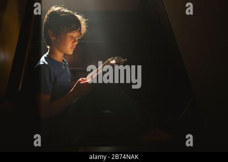 A boy sits on a darkened staircase in a bright beam of light reading Stock Photo