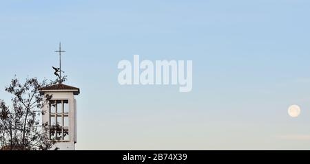 Angel figure with wings on the bell tower of a church at dawn that seems to be playing the trumpet to the full moon Stock Photo