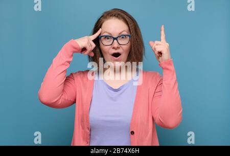 Caucasian young woman pointing up with her finger having great idea. Studio shot on blue wall. Stock Photo