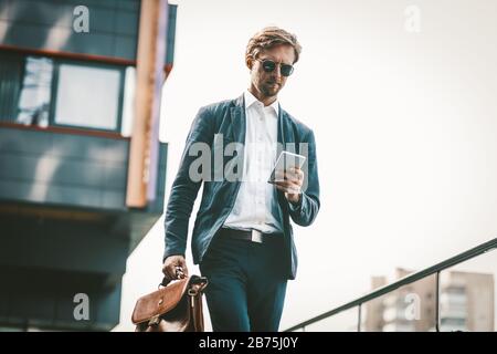 Young white colar or businessman walks outdoors with urban background. Handsome man in a blue sute texting on the phone and briefcase in the other han Stock Photo