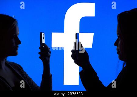 The silhouettes of two women with a smartphone in their hands are shown in front of a Facebook logo, on 12.01.2016. Illustration to Facebook friends. [automated translation] Stock Photo