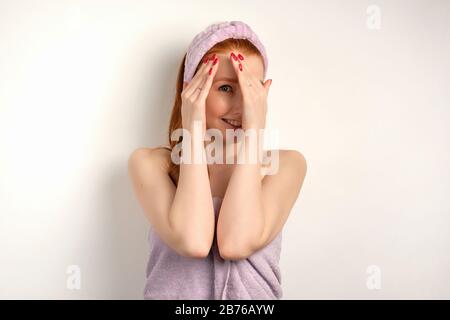 A red-haired girl with clean skin in a towel and a bandage on her head and hiding behind palms looks with one eye into the camera Stock Photo