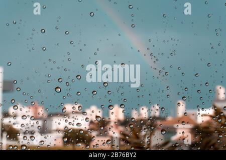 A rainbow over some buildings behind a glass windows with water drops. Point of view, from inside to outside. Stock Photo