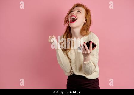 Beautiful red-haired girl with red lipstick screams joyfully up while standing on a pink background with a phone in her hands Stock Photo