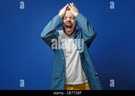 Handsome man is very upset, he holds hands on head and screams loudly, on a blue background. Mental health concept. Stock Photo