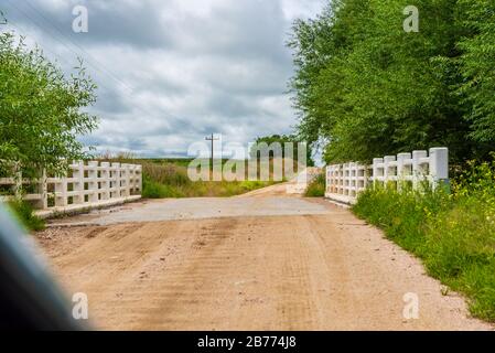 Small bridge on a dirt road with white fence next to it some trees and a clouded sky, electricity posts and plantations, seen from a car Stock Photo