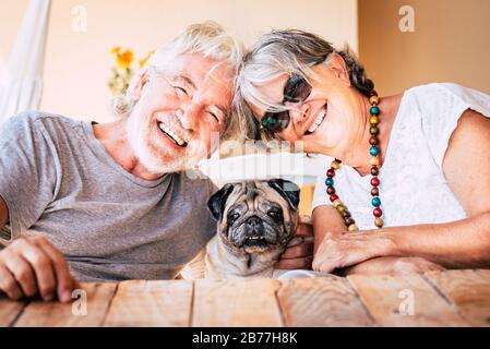 Happy couple of senior caucasian people have fun and smile with funny nice old dog pug in the middle sitting on the table together with them - retired Stock Photo