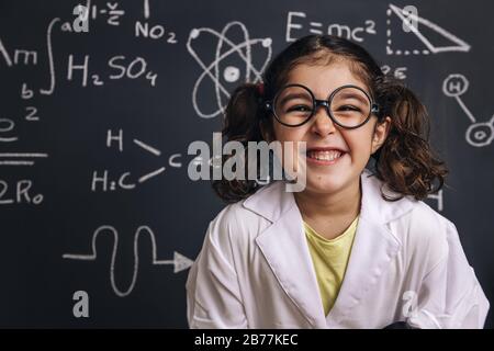 funny little girl science student with glasses in lab coat on school blackboard background with hand drawings science formula pattern, back to school Stock Photo