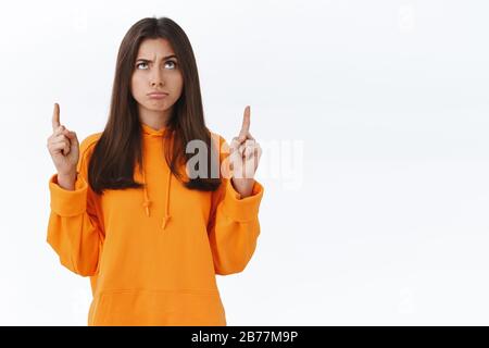 Perplexed and unsure attractive young woman in orange hoodie having doubts, consider wether product good or bad, grimacing puzzled and looking Stock Photo