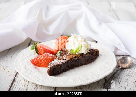 A slice of sticky chocolate mud cake, brownie or traditional Swedish kladdkaka on a white wooden table with a white napkin in the background. The cake