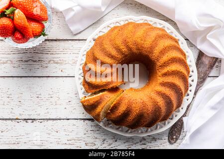 A white wooden table with a bundt cake , kugelhupf or sockerkaka. With a bowl of strawberries, a white napkin and a vintage cake cutter. Seen from abo Stock Photo