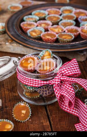 Swedish christmas butterscotch candy knäck in a glass jar, with a ribbon, on a rustic wooden table background. The candy is wrapped in small coloured Stock Photo