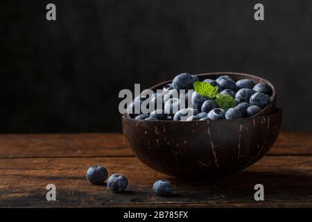 Blueberries or huckleberries and mint leaf in a coconut bowl. The bowl is on a rustic wooden table with a gray background. There are some blueberries Stock Photo