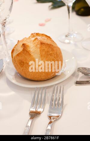 A bread bun on a plate on an elegant table with some cutlery and glass over a white cloth. Stock Photo