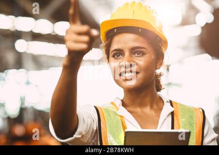 American lady worker in factory maintenance engineer happy working wearing safety uniform and helmet hand pointing and smiling for future of worker we Stock Photo