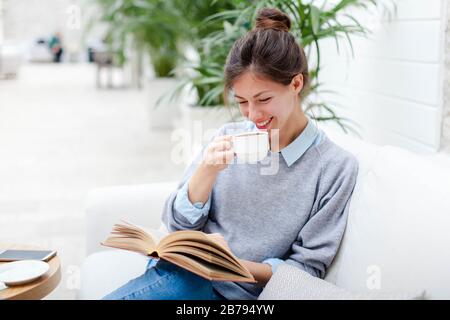 Young woman is drinking coffee and reading book. Girl is smiling, relaxation and sitting on sofa in cozy modern cafe