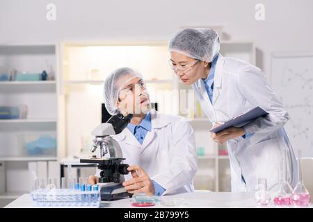 Two young intercultural scientists in whitecoats consulting in lab while working over new vaccine and studying viruses Stock Photo