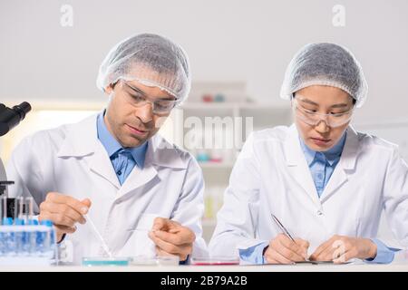 Young male chemist studying samples of chemical substances while his assistant near by making notes during scientific research in lab Stock Photo