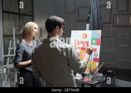 Teacher of painting consulting one of his students while helping her with unfinished picture on canvas in front of easel Stock Photo