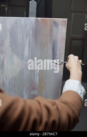 Hand of young female artist using special tool for painting while applying silver grey paint on paper during work in studio Stock Photo