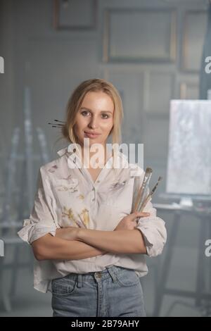 Pretty young blond woman in white shirt and grey jeans crossing arms by chest in front of camera with easel on background Stock Photo