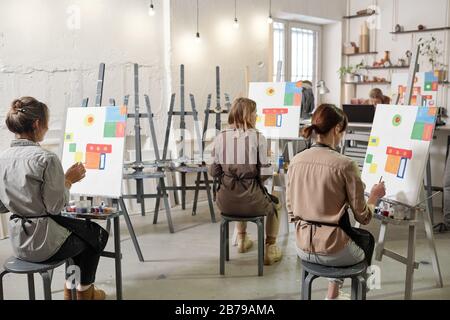 Rear view of several female students of painting class sitting on chairs in front of easels in classroom or studio and working Stock Photo