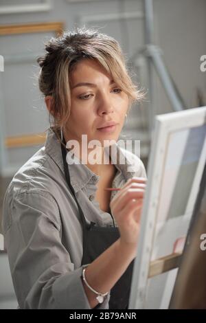 Pretty young female student in apron sitting in front of easel while working over unfinished painting at lesson in school of arts Stock Photo