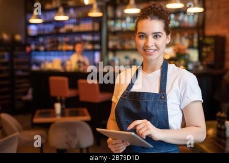 Young smiling waitress in apron and t-shirt standing in front of camera while using touchpad and meeting guests in cafe Stock Photo