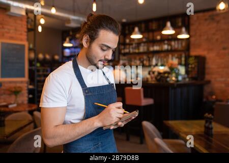 Young waiter in blue apron and white t-shirt writing down order of one of clients in notepad while standing in cafe or restaurant Stock Photo
