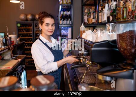 Young waitress of cafe looking at you while standing by coffee machine and preparing drinks for clients at work Stock Photo