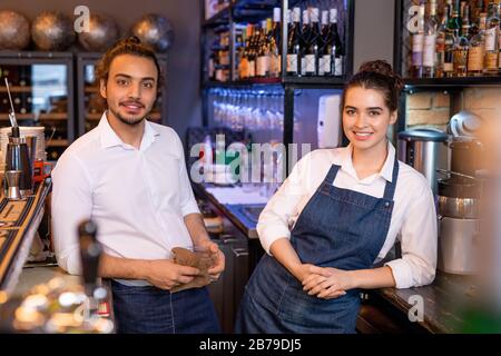 Two young restful workers of cafe standing next to each other in front of camera on background of shelf with wine assortment Stock Photo