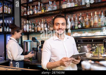 Young cheerful waiter with touchpad standing in front of camera while looking through online orders by workplace Stock Photo