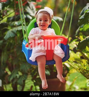 Baby girl in white dress sitting on swing in park background Stock Photo