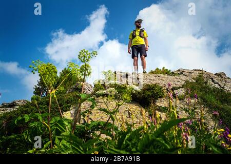 Athlete runner stands on the edge of a cliff in the mountains. Man in a yellow T-shirt and black shorts is training in outdoors. Trail running Stock Photo