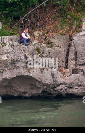 Guilin, China - May 10, 2010: Along Li River. Lone male fisherman sitting on gray rocks over greenish water with brown and green forest foliage in bac Stock Photo