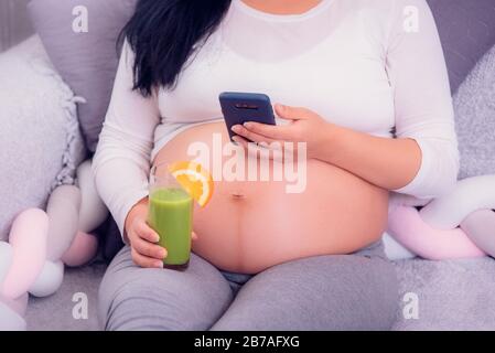 Pregnant woman using her smartphone while relaxing on the bed, drinking healthy green juice and exposing belly. Stock Photo