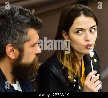 Girl looks at bearded guy holding fork in mouth on brown terrace background. Food and lunch time concept. Woman and man with surprised faces have date at cafe. Couple in love takes a bite during break Stock Photo