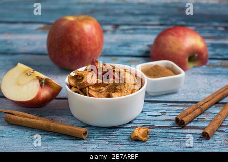 Sun-dried apples in a white bowl, with dried cinnamon bark strips beside it; pieces of an apple and cinnamon bark powder are in a blurry background. Stock Photo