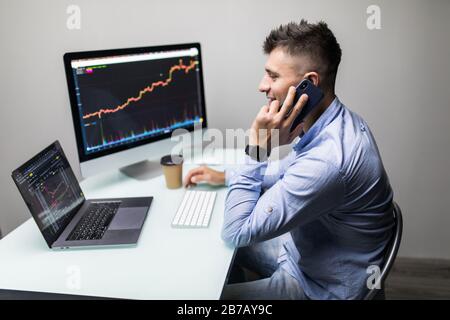 Smiling young male online stock trader talking on smartphone with client while using laptop at desk Stock Photo