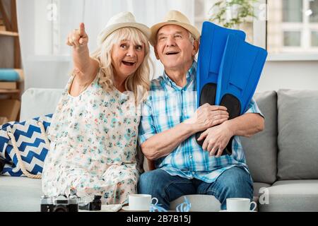 Senior man and woman sitting in travel agency sitting on sofa wearing beach hats husband holding flippers while wife pointing up smiling surprised Stock Photo