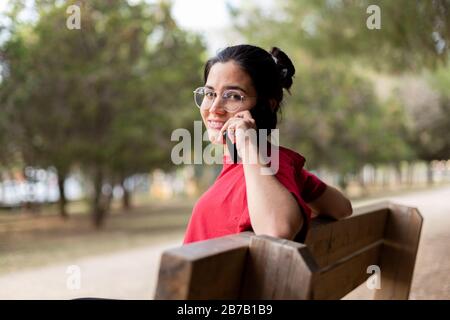 Young attractive woman talking through the phone and smiling in a park, sit down in a bench Stock Photo