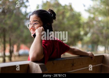 Young attractive woman talking through the phone and smiling in a park, sit down in a bench Stock Photo