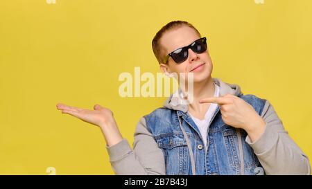 Stylish Caucasian man in a jeans and sunglasses on a yellow background. Copy space. Close up. Finger shows abstractly on the palm object of the hand. Stock Photo