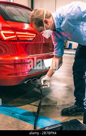 A young caucasian technician doing an emission control in a security inspection of a vehicle protected with a mask and gloves to prevent the spread of Stock Photo