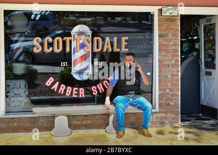 A smiling, bearded, trendy man on cell phone sits on bench in front of the Scottsdale Barber Shop, in Old town, Scottsdale, AZ, USA Stock Photo