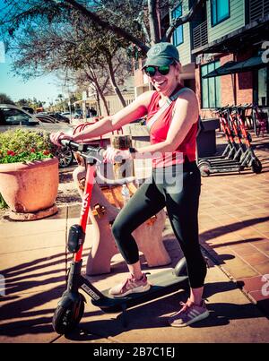 A woman smiling while taking a break on her rented, stand up electric scooter in old town Scottsdale, AZ, USA Stock Photo