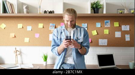 Smiling millennial man standing at home office workspace using cell phone. Stock Photo