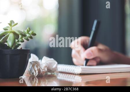 Closeup image of a hand working and writing down on a white blank notebook with screwed up papers on table Stock Photo