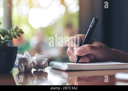 Closeup image of a hand working and writing down on a white blank notebook with screwed up papers on table Stock Photo