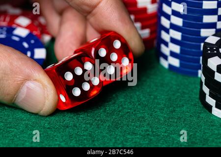 Macro of a male caucasian hand picking up 2 translucent red casino style dice with white pips surounded by red, blue and black betting chips, over a g Stock Photo